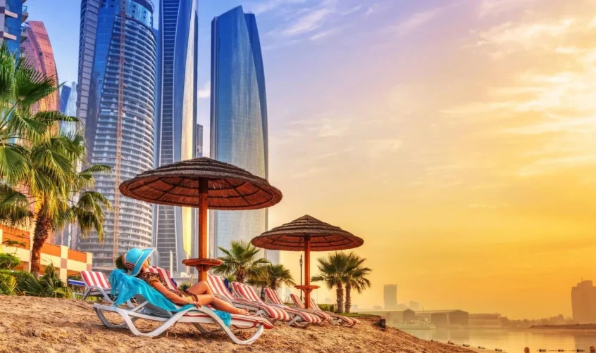 Eine Frau entspannt sich am Strand von Abu Dhabi mit Blick auf moderne Wolkenkratzer bei Sonnenuntergang.