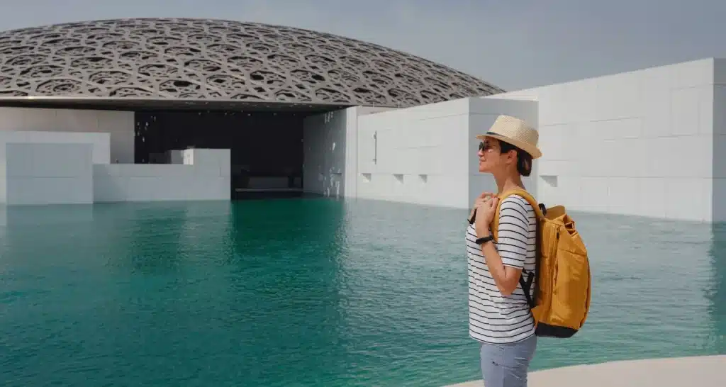 Ein Tourist mit einem Rucksack steht vor dem Louvre Abu Dhabi und blickt auf das Wasser.