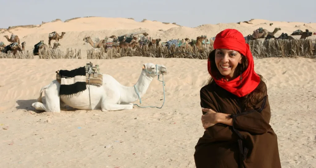 Frau mit traditionellem Kopftuch sitzt in der Wüste neben Kamelen