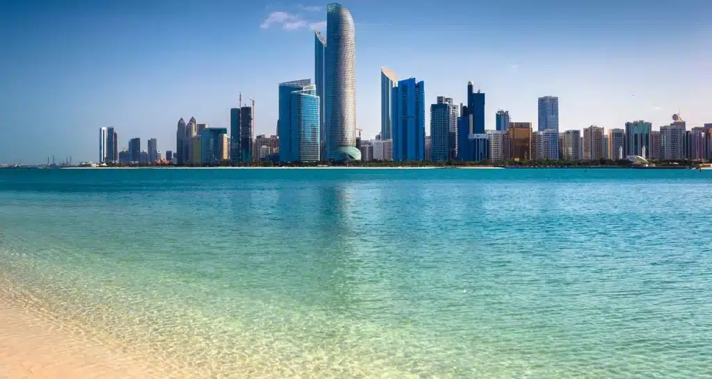 Blick auf die Skyline von Abu Dhabi vom Strand aus mit klarem türkisfarbenem Wasser und Spiegelung der Stadt.