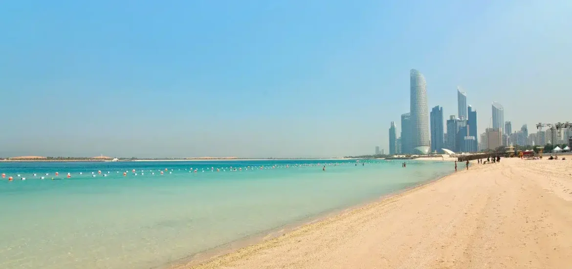 Die Küste des Corniche Beach mit klarem türkisfarbenem Wasser und der Skyline von Abu Dhabi im Hintergrund.