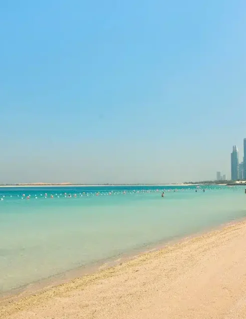 Die Küste des Corniche Beach mit klarem türkisfarbenem Wasser und der Skyline von Abu Dhabi im Hintergrund.