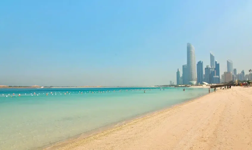 Die Küste des Corniche Beach mit klarem türkisfarbenem Wasser und der Skyline von Abu Dhabi im Hintergrund.